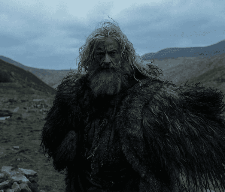 Homme barbu aux longs cheveux gris, vêtu d’une peau sombre, dans un paysage montagneux nuageux