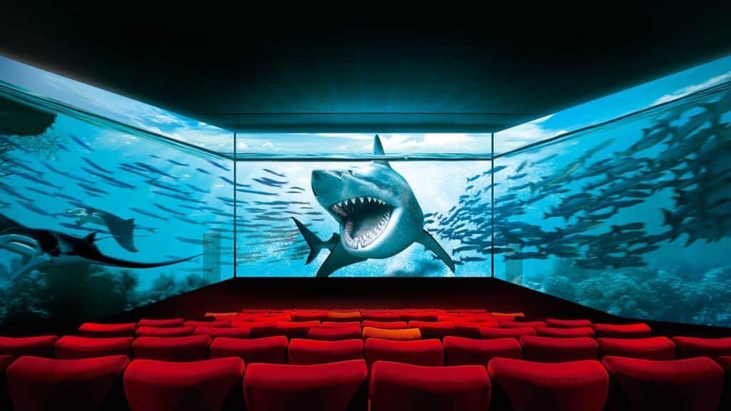Un requin à gueule ouverte dans un aquarium, vu depuis une salle de cinéma aux sièges rouges.