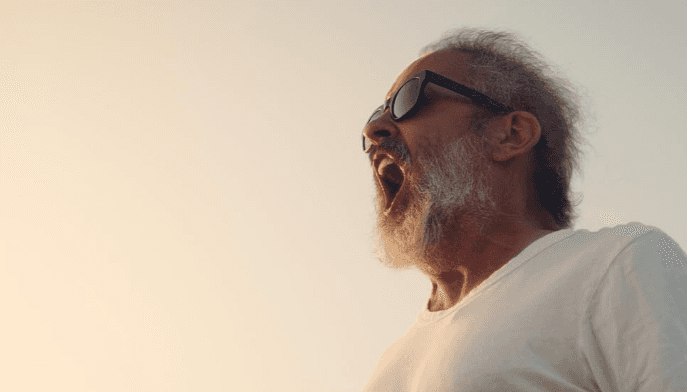 Homme barbu portant des lunettes noires, criant face au ciel, sur fond lumineux.