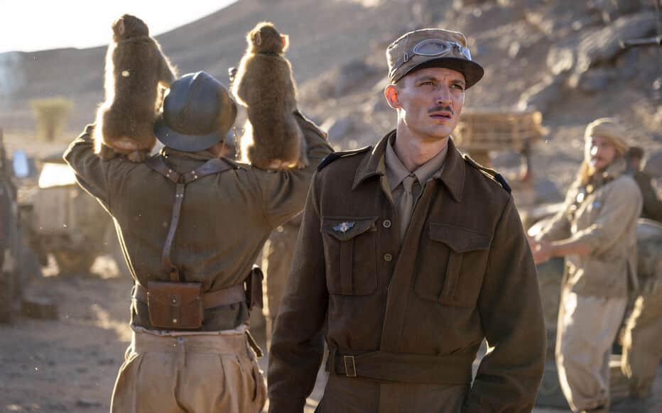 Un soldat au premier plan, devant un militaire portant deux singes sur ses épaules dans un camp montagneux