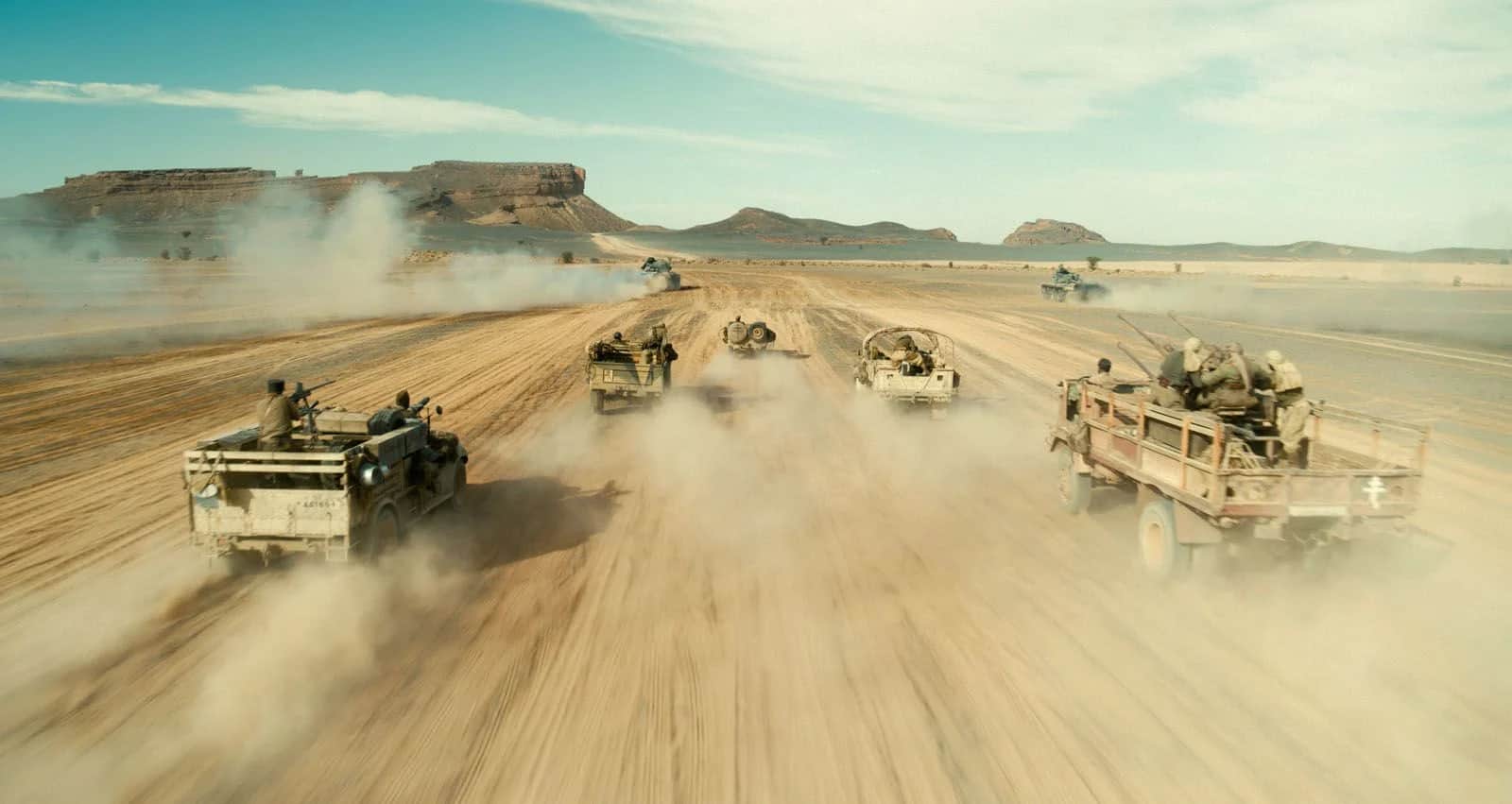 Un convoi de véhicules tout-terrain traverse un désert poussiéreux bordé de plateaux rocheux.