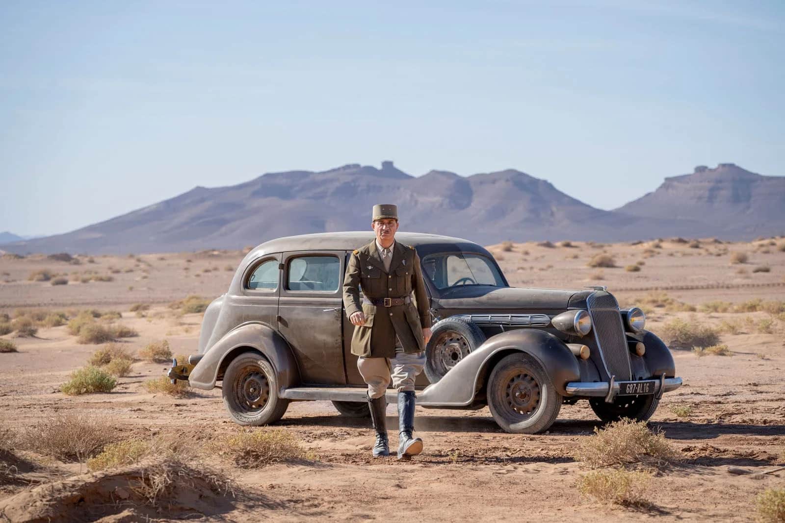 Un homme en uniforme militaire marche devant une voiture ancienne dans un désert montagneux.