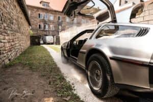 Voiture de sport argentée aux portes papillon ouvertes, garée dans une ruelle bordée de murs en pierre.