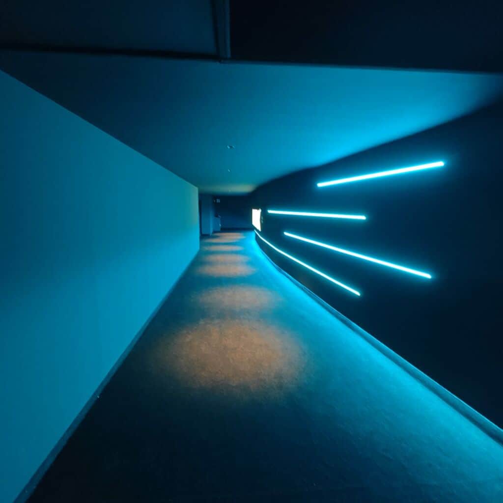 Couloir sombre éclairé par des néons bleus, avec un écran lumineux au fond.