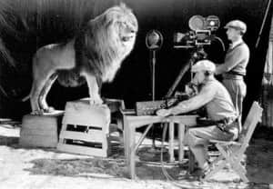 Un lion debout sur une caisse, filmé par deux hommes avec une caméra sur un plateau de tournage