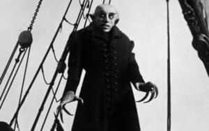 Un homme chauve au visage pâle, vêtu de noir, se tient sur le pont d’un navire.