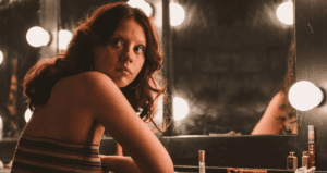 Femme aux cheveux roux assise devant un miroir de loge éclairé de lampes rondes.