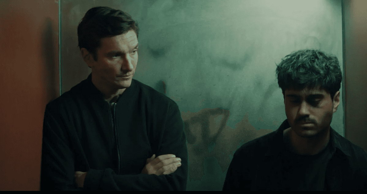 Deux hommes se tiennent dans un ascenseur sombre, l’un les bras croisés et l’autre regardant vers le bas.