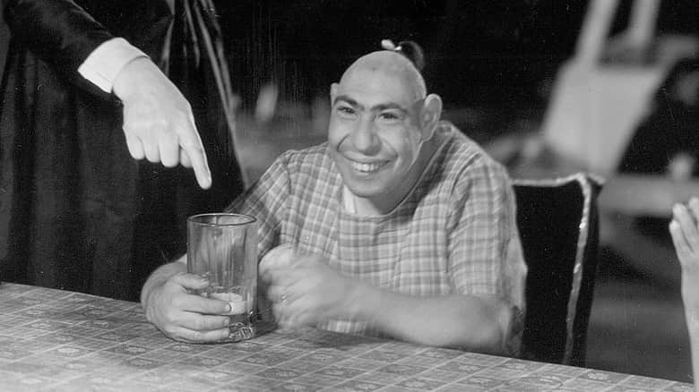 Homme souriant tenant un verre à table, tandis qu’une personne pointe le verre dans une scène en noir et blanc
