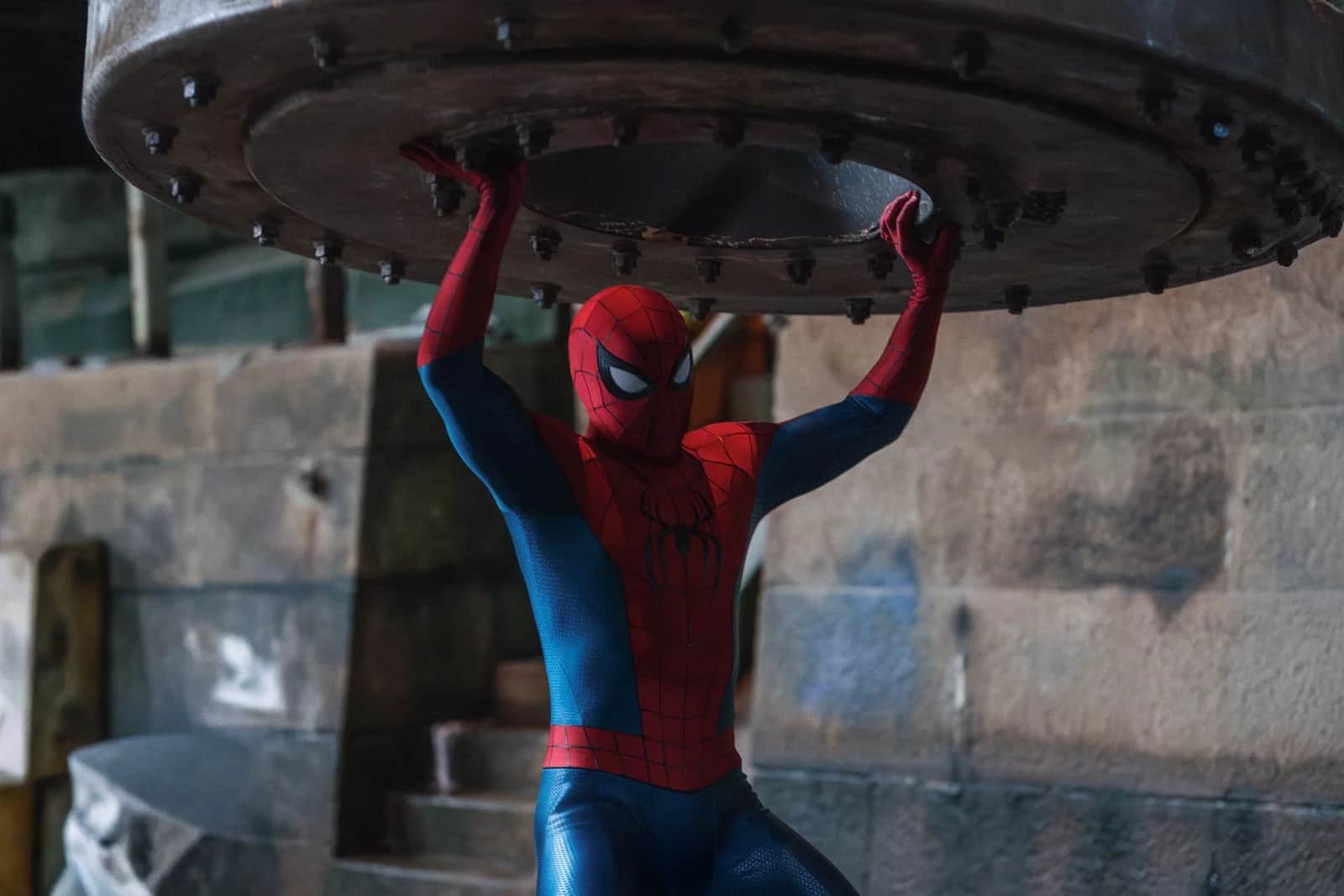 Homme en costume de Spider-Man soutenant une lourde structure métallique au-dessus de lui.