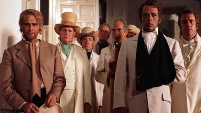 Un groupe de huit hommes en costumes d'époque, certains portant des chapeaux, se tenant dans un intérieur élégant.