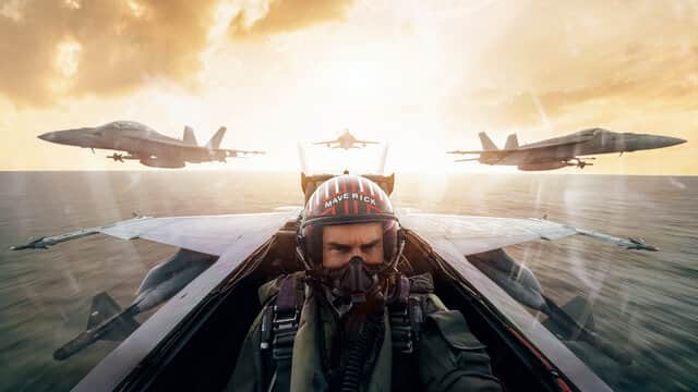 Un pilote dans un cockpit avec deux avions de chasse en formation derrière, survolant la mer au coucher du soleil.
