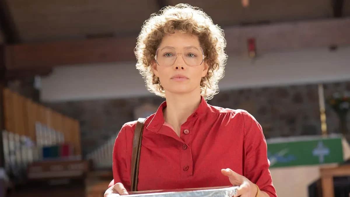 Femme aux cheveux bouclés et lunettes, en chemise rouge, tenant un carton dans un hall.