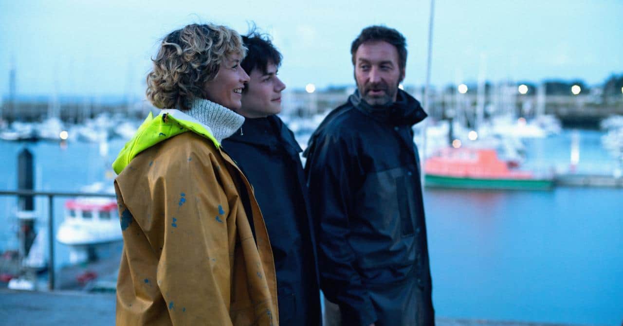 Trois personnes en vêtements de pluie discutent sur un quai, avec un port et des bateaux en arrière-plan.