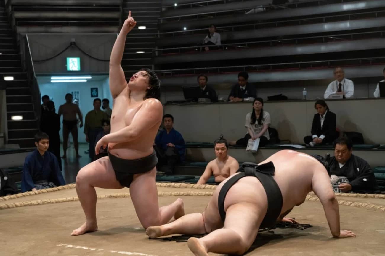 Deux lutteurs de sumo dans un dohyō, l’un levant un doigt tandis que l’autre est au sol, sous le regard du public.