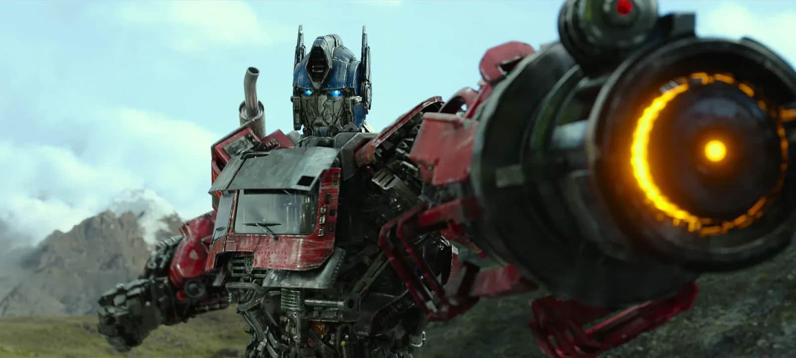 Robot rouge et bleu tenant un imposant canon, devant des montagnes sous un ciel nuageux