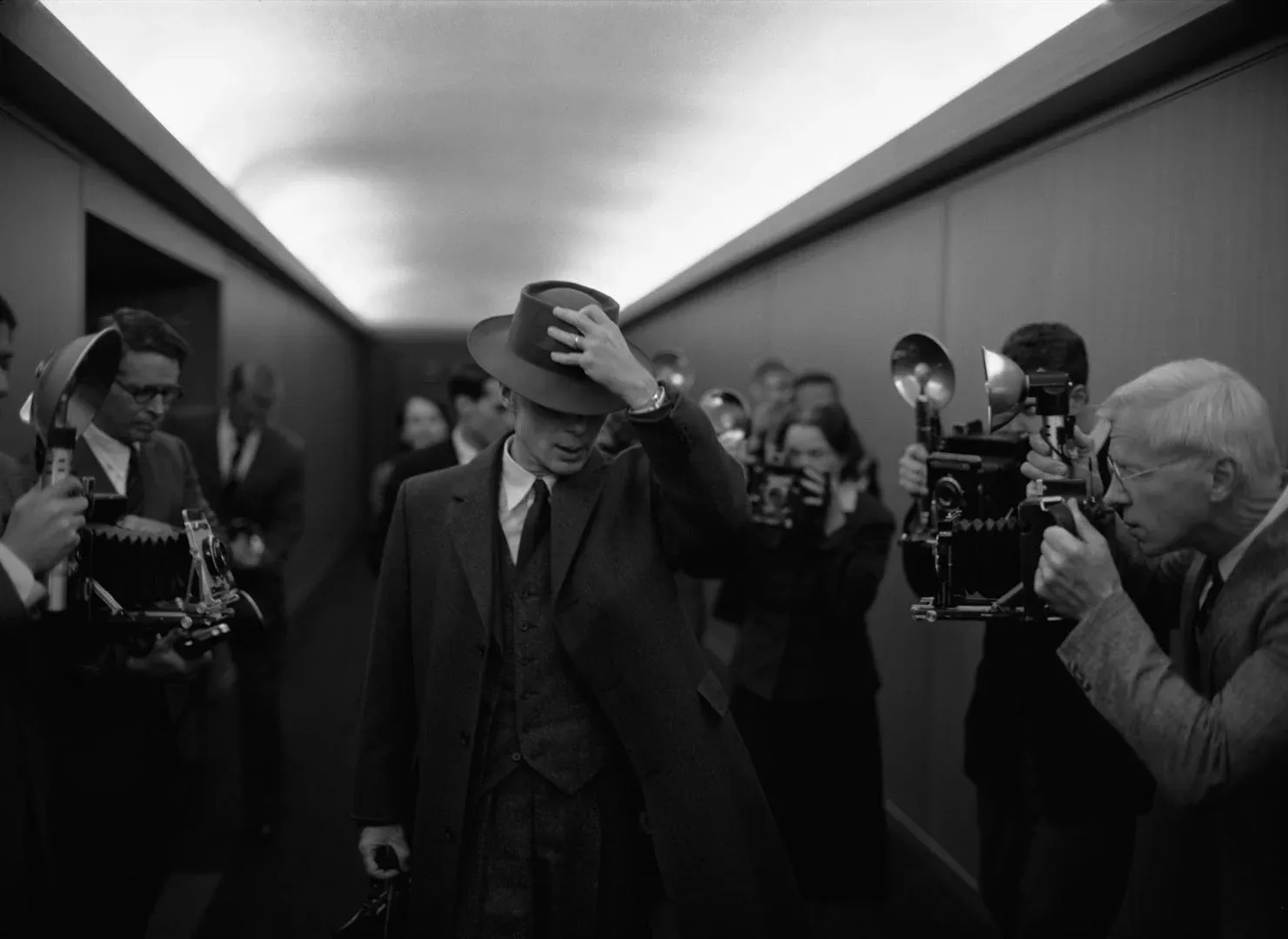 Un homme en costume et chapeau avance dans un couloir, entouré de photographes munis d'appareils anciens