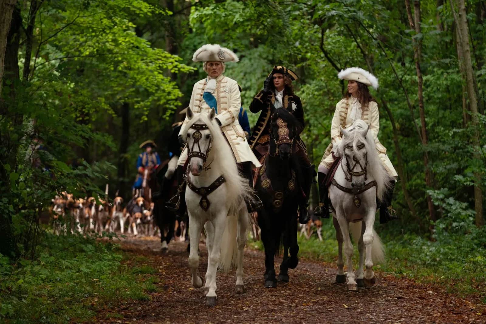 Trois cavaliers en costumes d’époque avancent à cheval sur un sentier forestier, suivis d’un groupe de personnes.