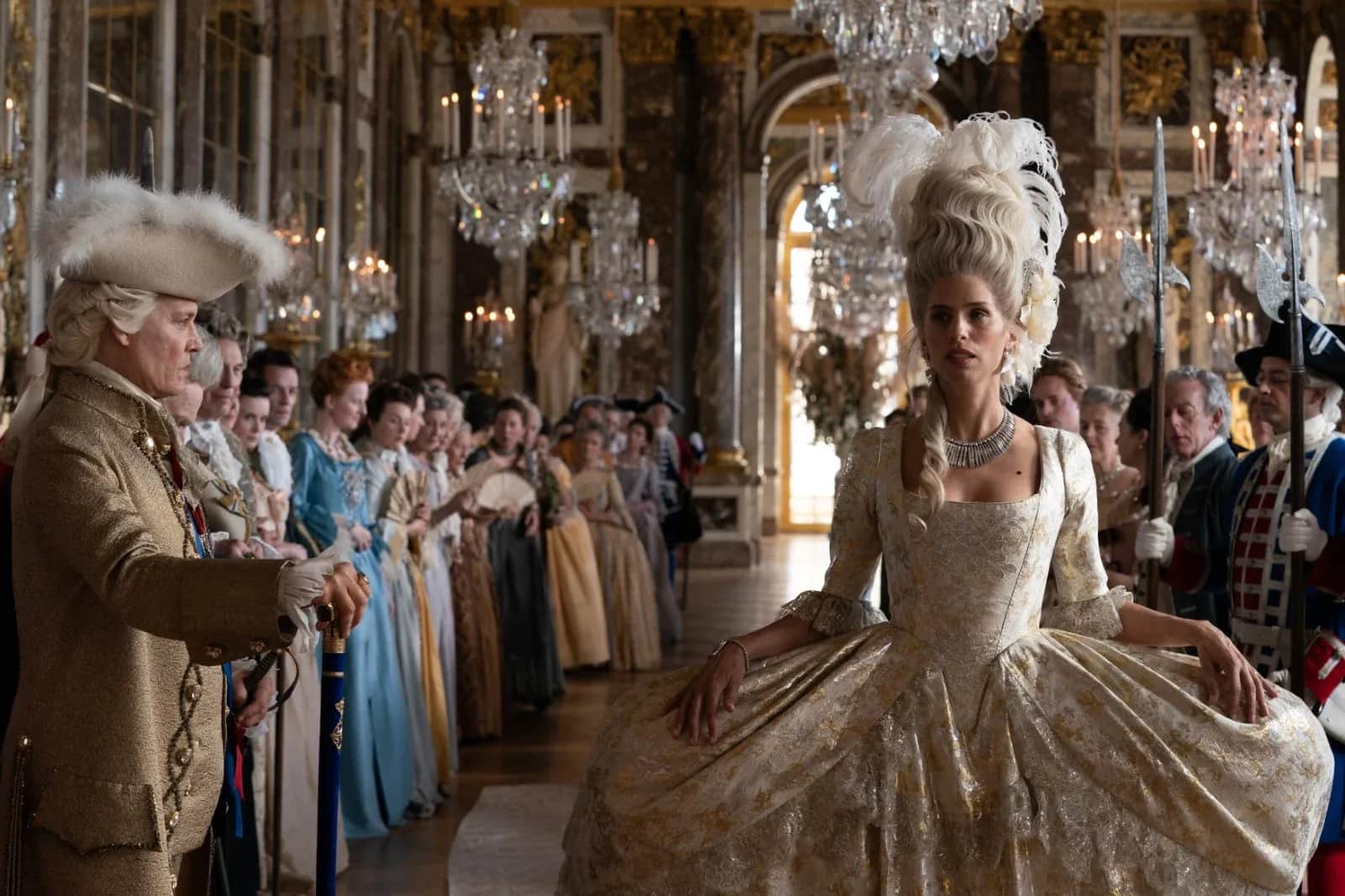 Une femme en robe blanche et coiffure monumentale avance dans une galerie fastueuse, entourée d’une foule costumée
