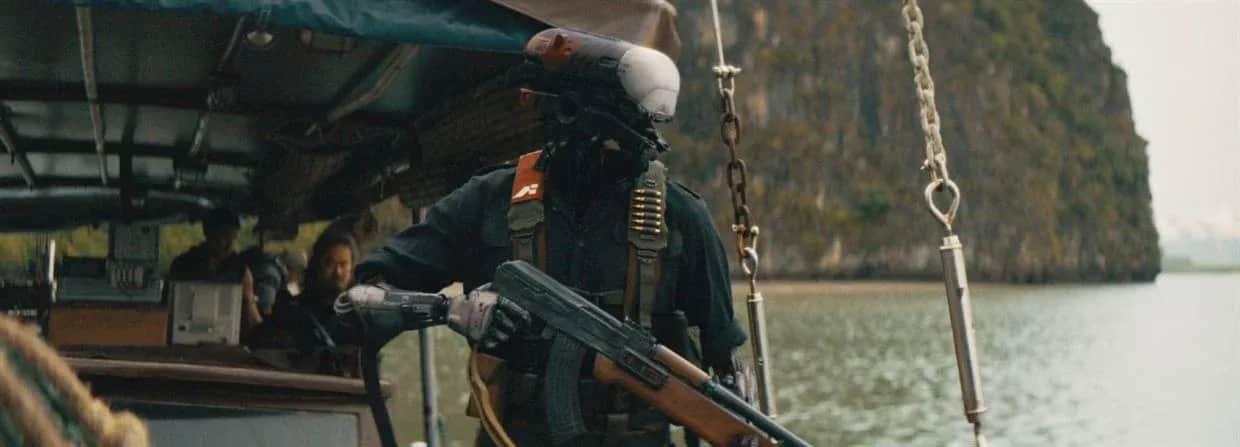 Un homme masqué armé d’un fusil à bord d’un bateau, devant une falaise boisée.