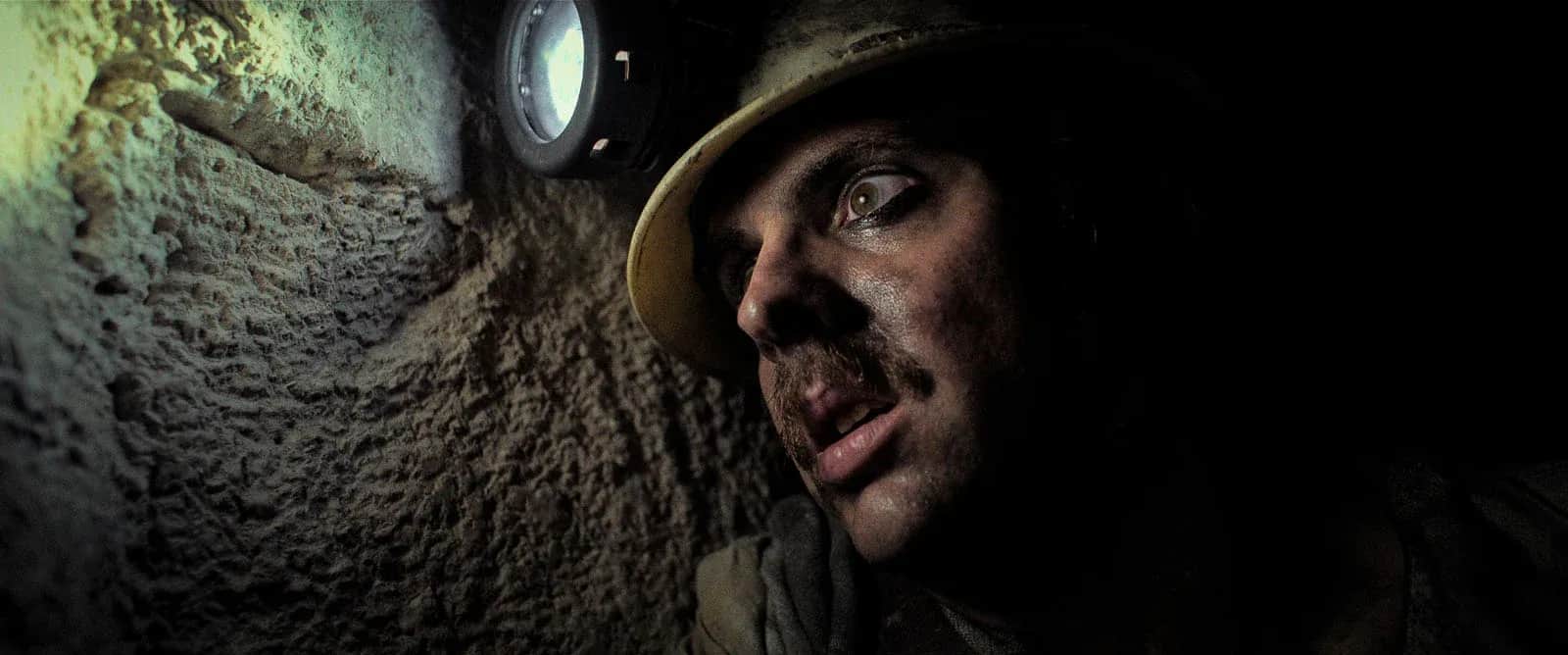 Un homme moustachu portant un casque avec lampe, dans un tunnel rocheux sombre.