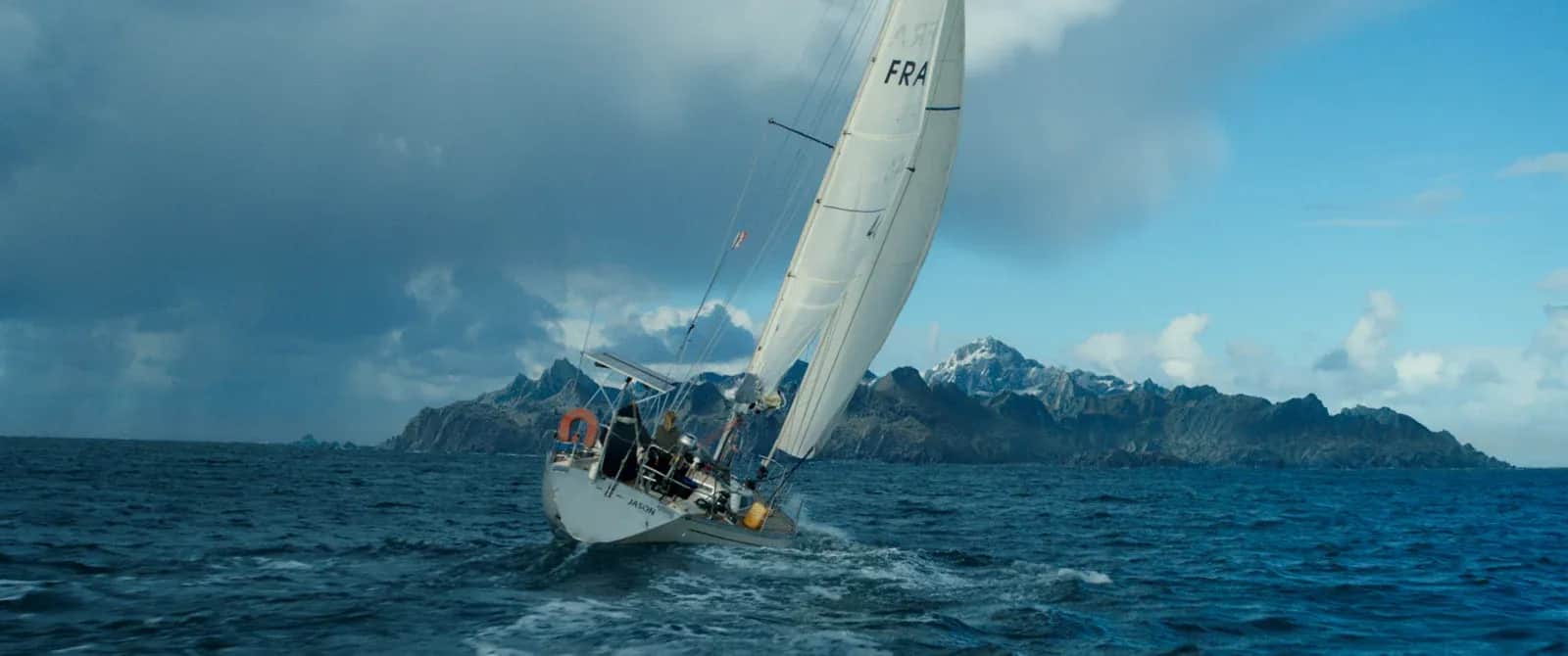 Voilier incliné sur une mer agitée, avec des montagnes rocheuses en arrière-plan.