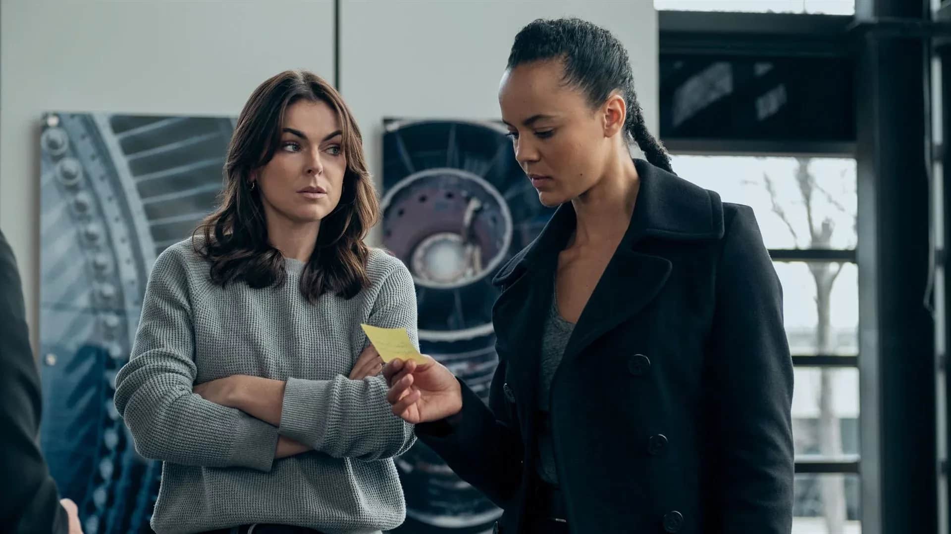 Deux femmes, dont une tient un papier jaune, se tiennent dans un intérieur industriel.