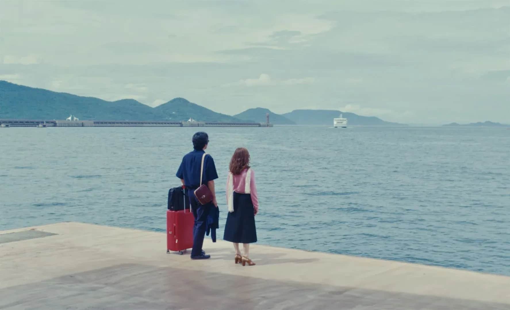 Deux personnes avec des bagages se tiennent sur un quai face à la mer, avec des montagnes et un bateau au loin.