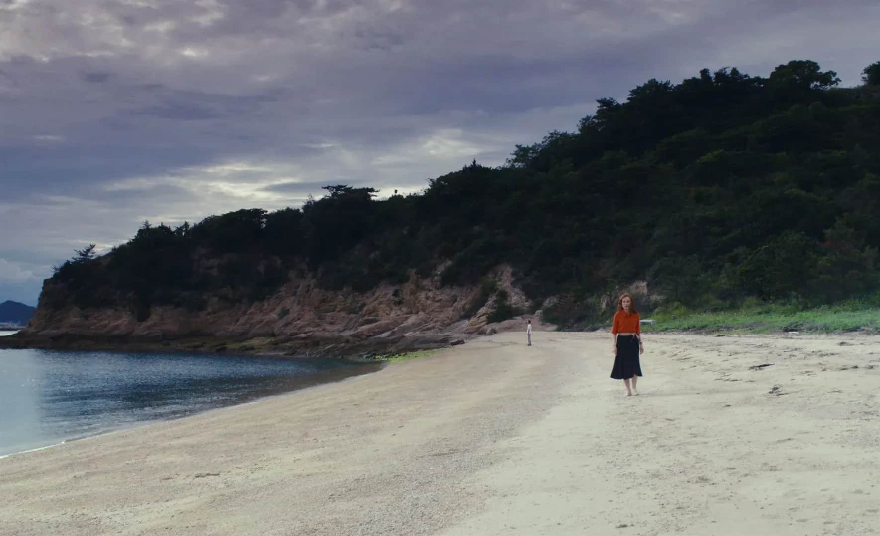 Femme en jupe marchant sur une plage déserte, avec une personne au loin et une colline boisée sous un ciel nuageux