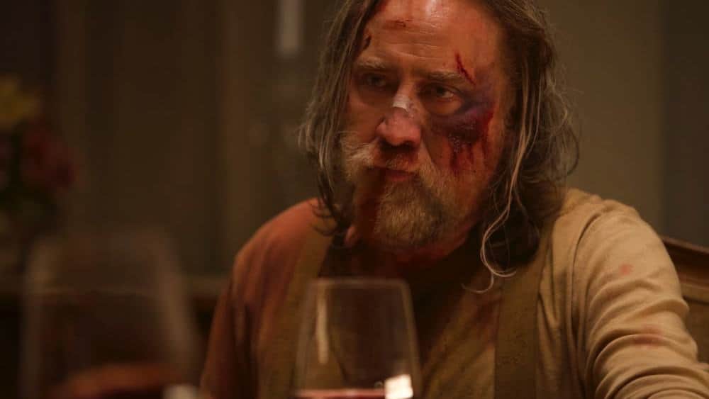 Homme barbu aux cheveux longs, le visage ensanglanté, assis à table devant un verre.