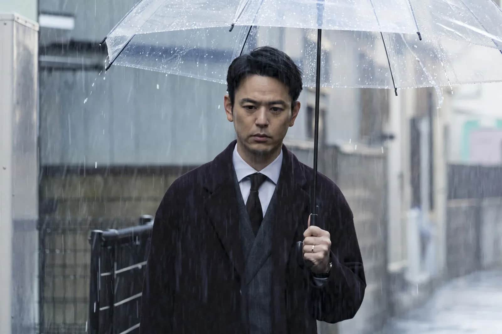 Homme en costume tenant un parapluie transparent sous la pluie, dans une rue urbaine.