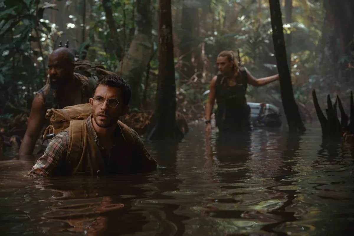 Un groupe de trois personnes avance dans une eau sombre au milieu d’une jungle.