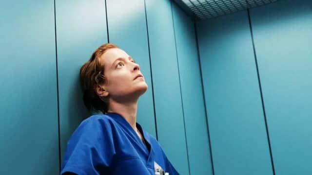 Une personne en blouse bleue regarde vers le haut dans un ascenseur aux parois bleues.