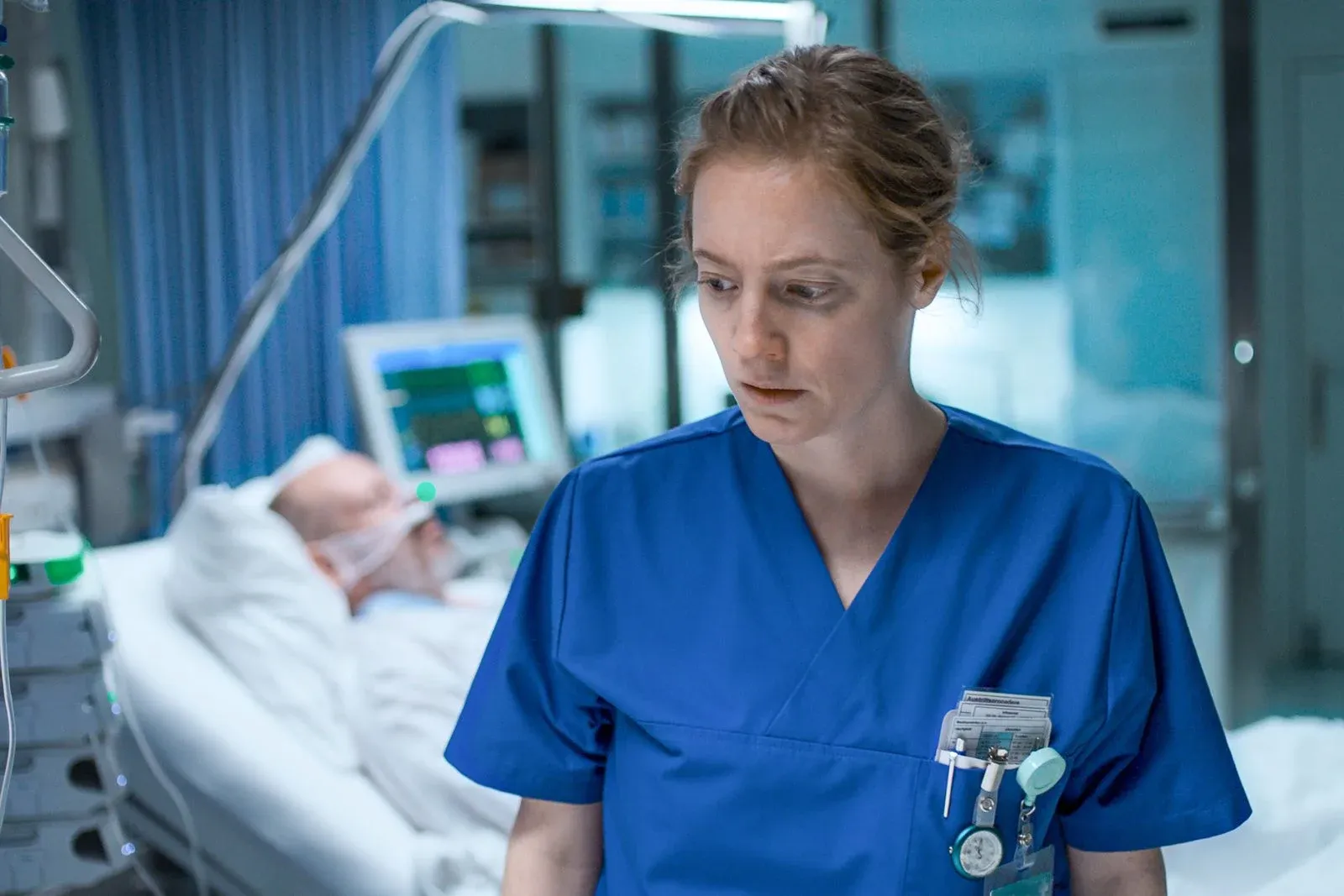 Infirmière en blouse bleue au chevet d’un patient hospitalisé, dans une chambre médicalisée