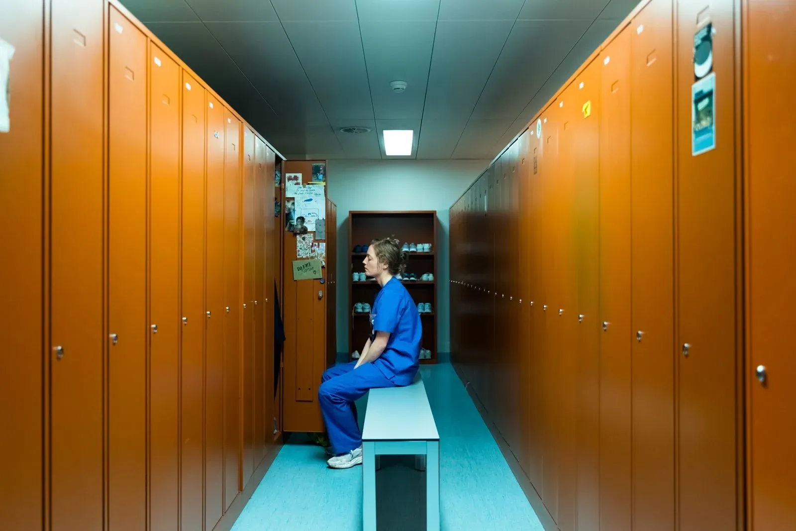 Une femme en tenue médicale bleue assise sur un banc entre des casiers orange.