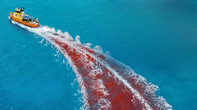 Un bateau jaune navigue sur une mer bleue, laissant une traînée rouge derrière lui.