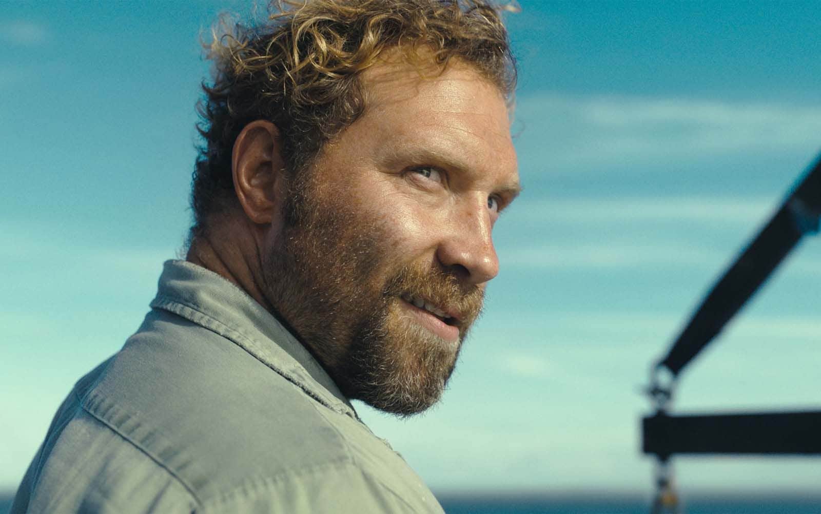 Homme barbu en veste claire, tournant la tête vers la caméra devant un ciel bleu et la mer.