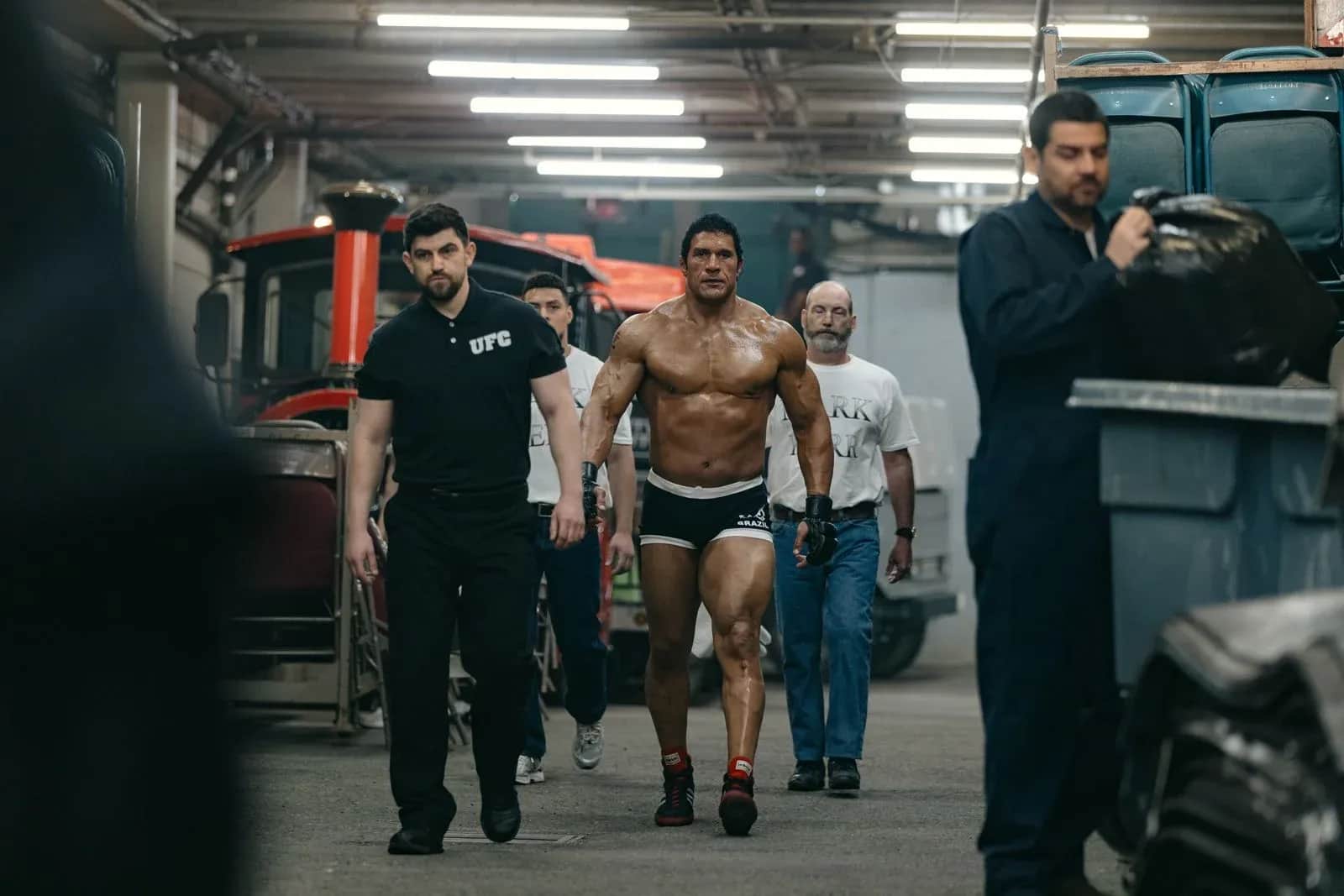 Un combattant torse nu avance avec un groupe de personnes dans un garage éclairé, entre des véhicules.