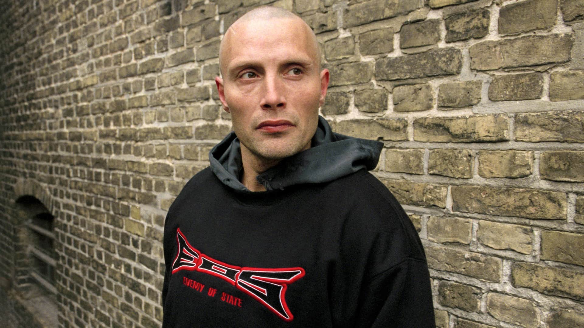 Homme chauve en sweat noir, debout devant un mur de briques.
