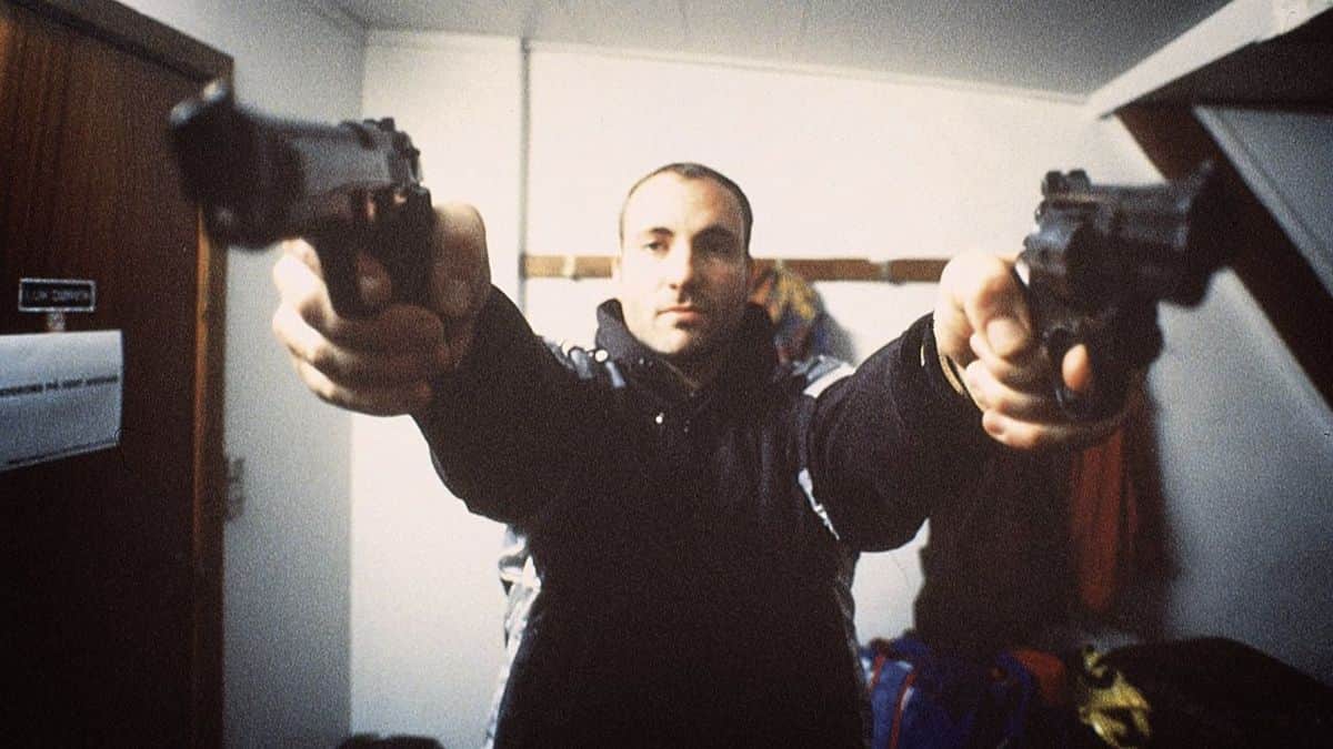 Homme braquant deux pistolets vers l’objectif dans une pièce.