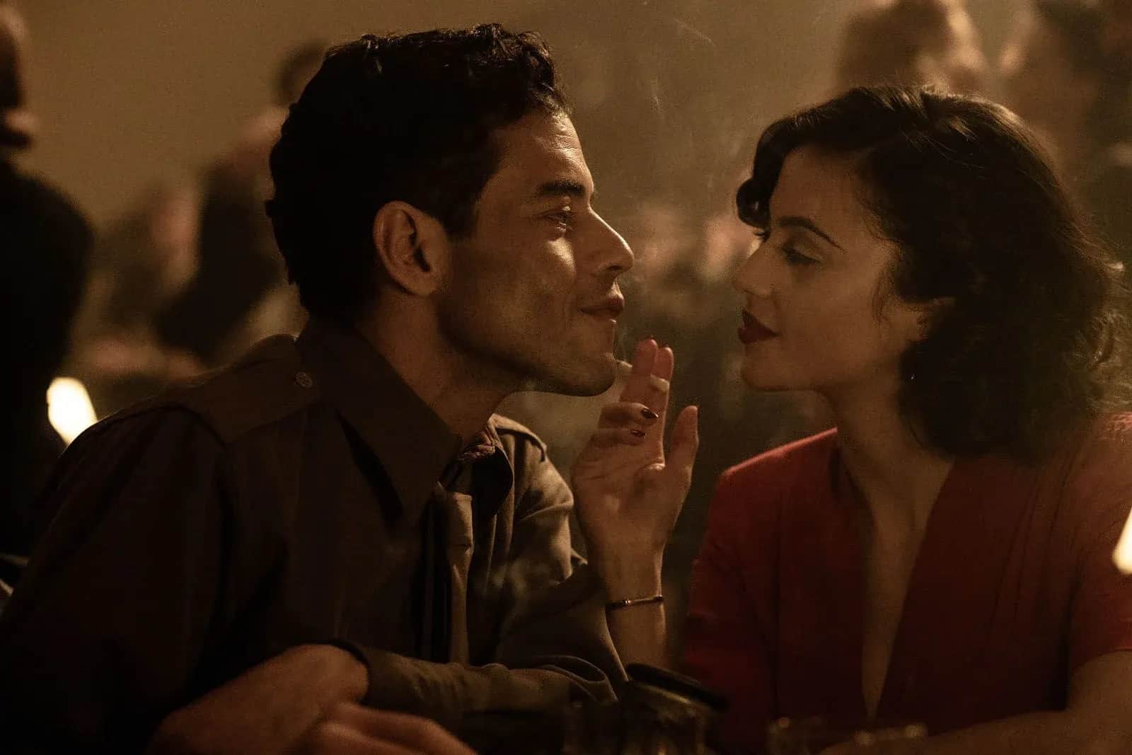 Un homme et une femme se font face, attablés dans une salle sombre, la femme levant la main entre eux.
