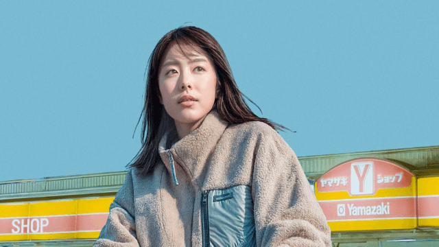 Une femme en manteau beige devant un magasin Yamazaki avec un ciel bleu en arrière-plan.