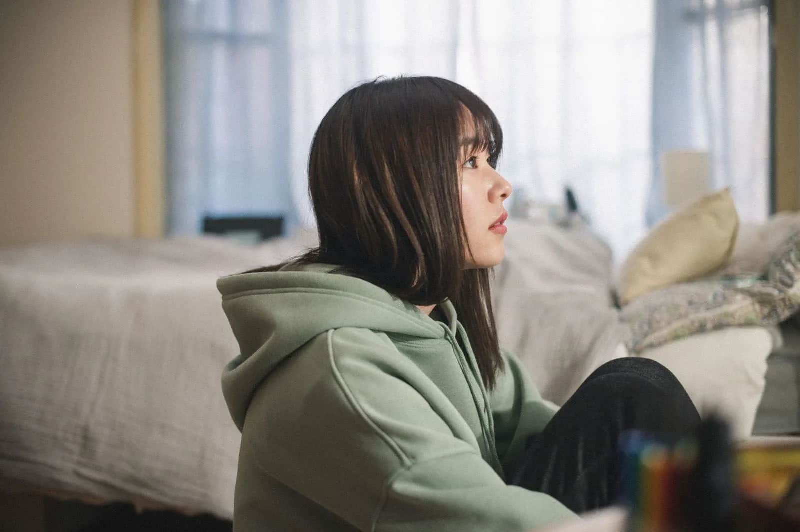 Jeune femme assise de profil, portant un sweat vert, dans une chambre près d’un lit.
