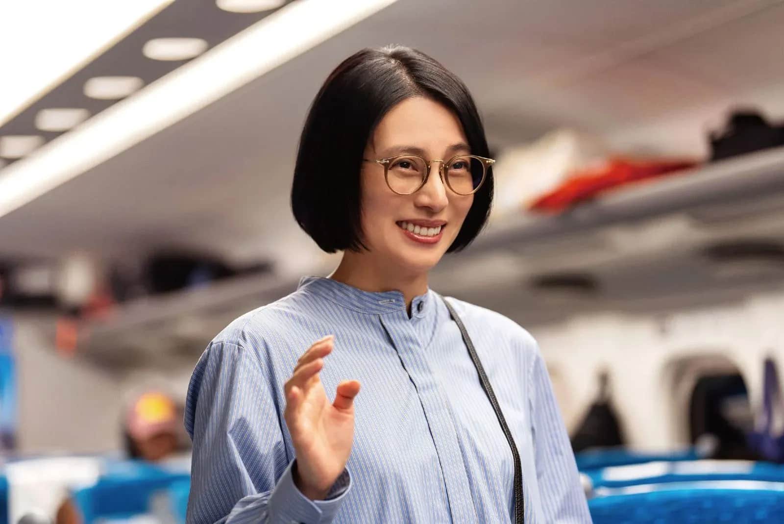 Femme souriante aux cheveux courts et lunettes, levant la main dans un avion.