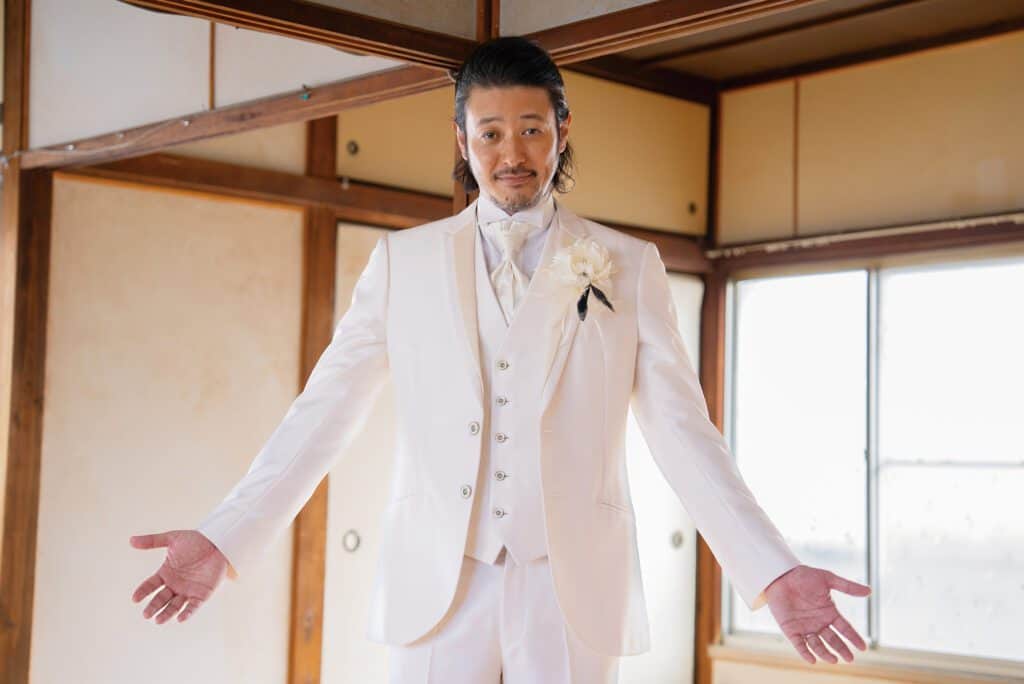 Homme en costume blanc, bras ouverts, dans une pièce traditionnelle japonaise.