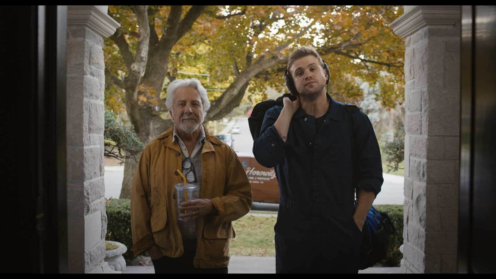 Deux hommes devant l’entrée d’une maison, l’un portant un casque audio et l’autre tenant une boisson.