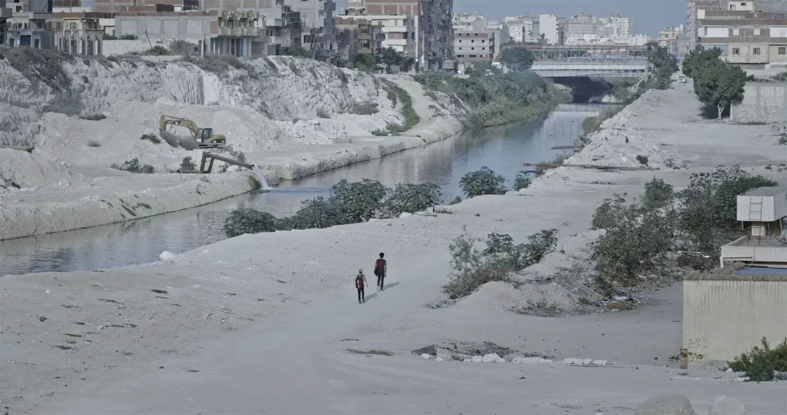 Deux personnes marchent dans un paysage aride, le long d’un canal bordé d’immeubles et de chantiers.