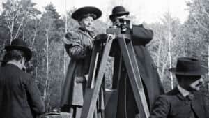Deux hommes en chapeaux observent une caméra sur trépied, entourés de deux autres hommes en forêt
