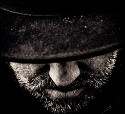 Visage d’un homme barbu portant un chapeau noir, vu en gros plan dans l’ombre