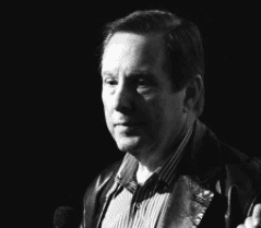 Homme en noir et blanc, regardant sur le côté, vêtu d’une veste en cuir et d’une chemise rayée.